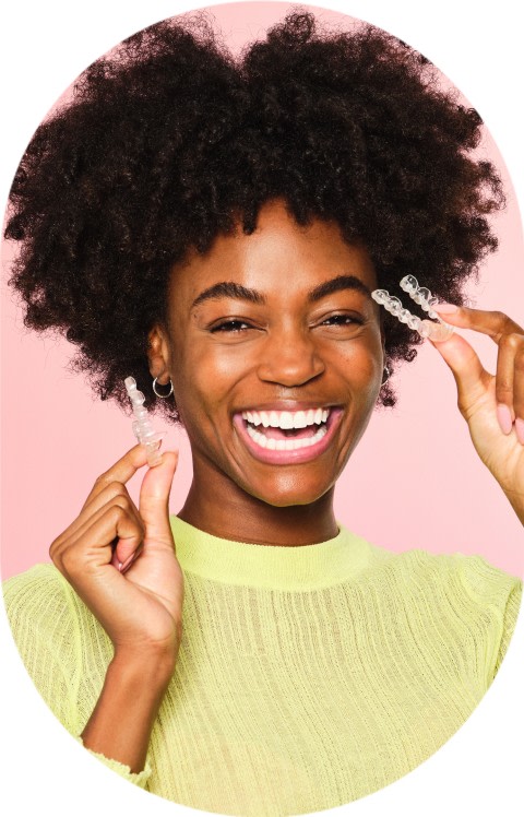 A woman with curly hair is smiling and holding clear aligners.
