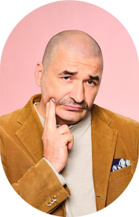 A man with a curled mustache is pointing at his cheek.