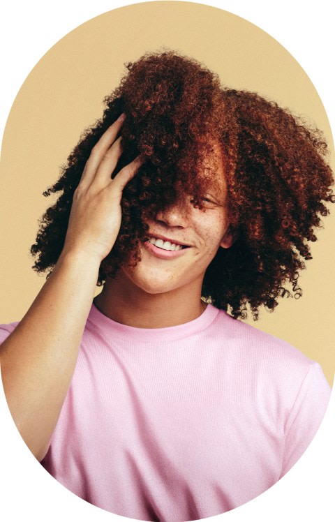A man with curly hair smiles with his hand on his hair.